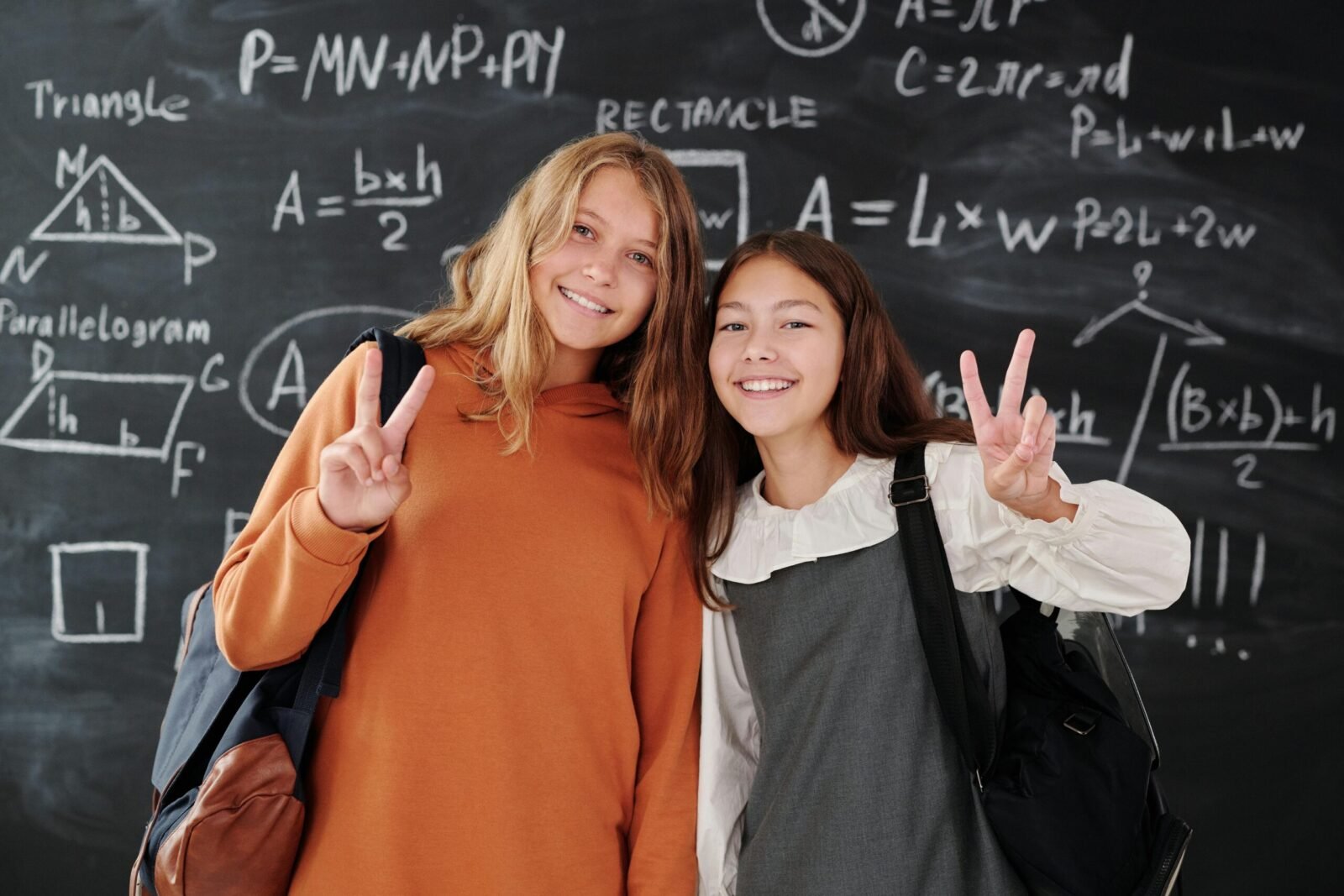 Two smiling students with backpacks in front of a math chalkboard in a classroom.